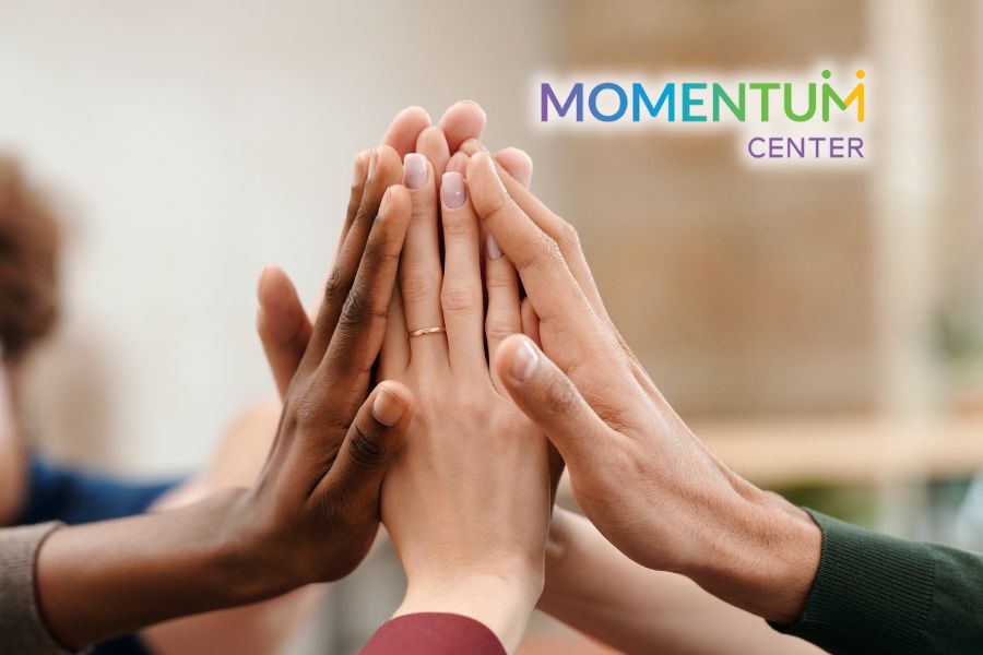 A diverse group of hands doing a group high five with a blurred out background and the Momentum Center logo in the upper right corner