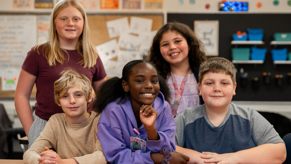 A group of five students - two boys and three girls - smiling while sitting at a classroom table.
