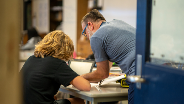 A look through a classroom door of a teacher helping a student at his desk.
