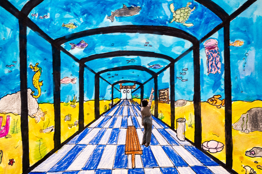 A student art piece made with crayon and watercolors, showcasing the use of perspective looking down an aquarium-like hallway with a student in the middle pointing at the sea life surrounding them through glass walls.