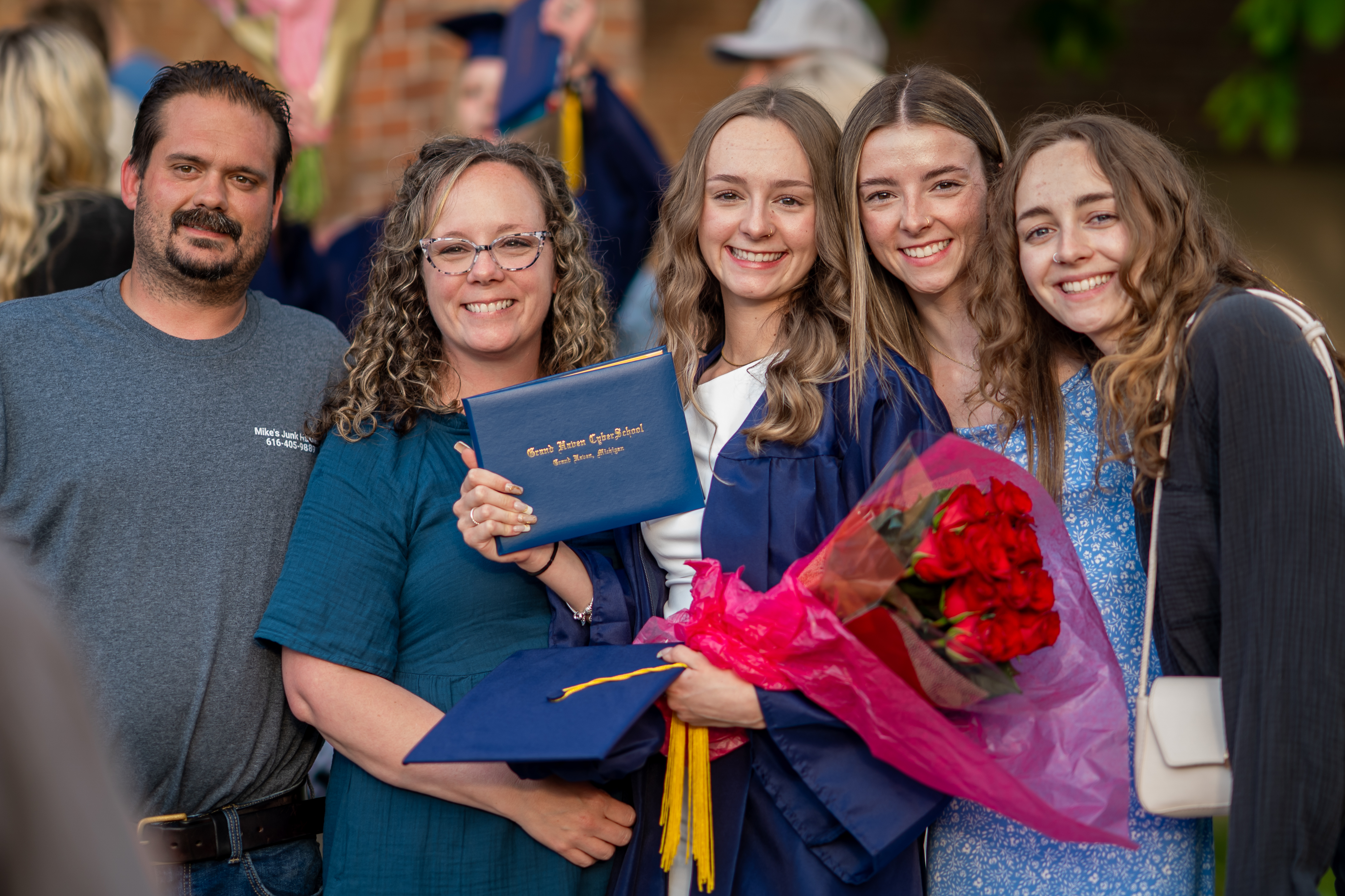 A family with a dad, mom, and three sisters with one of the sisters in the middle wearing her cap and gown from graduation and proudly displaying her new diploma and flowers.