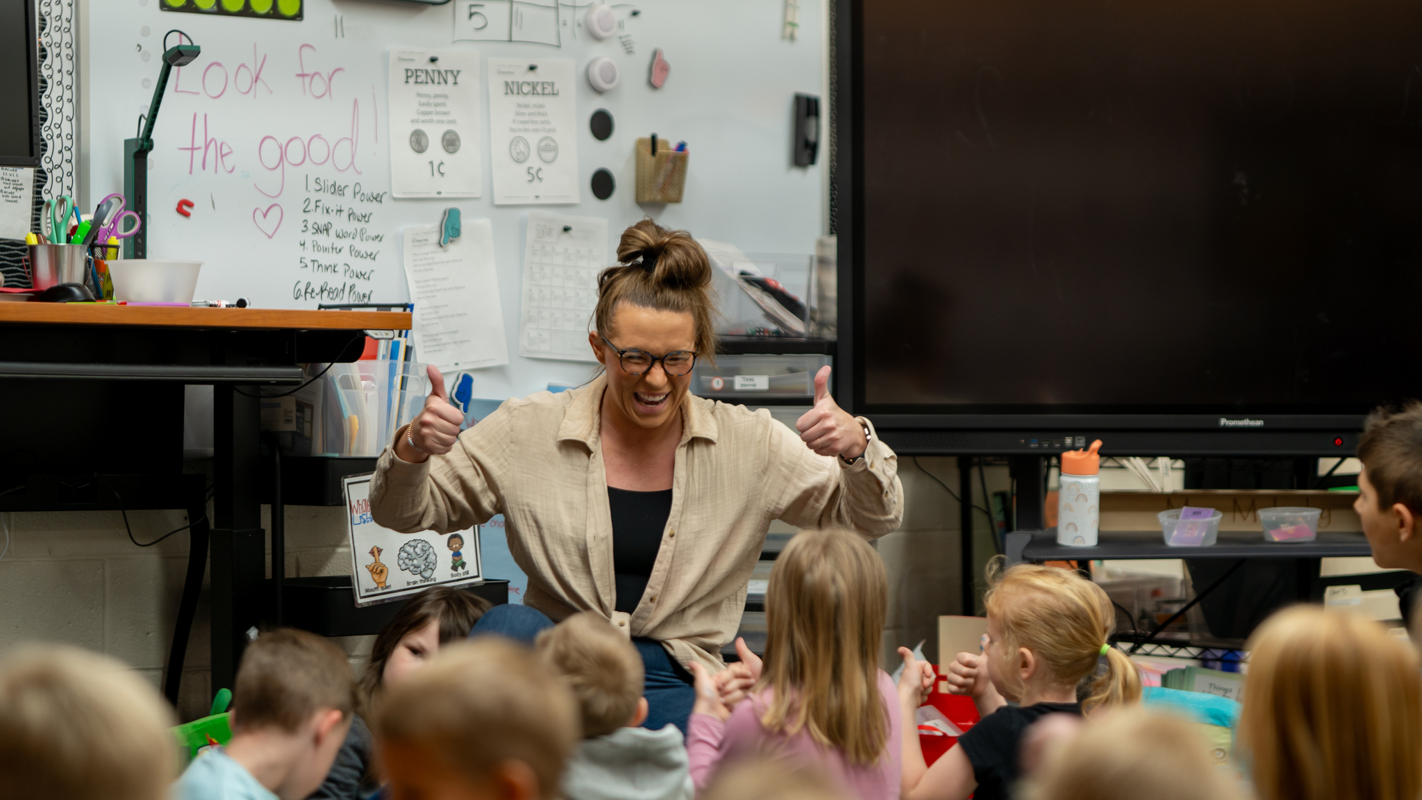 A teacher smiling and giving two thumbs up to a group of children