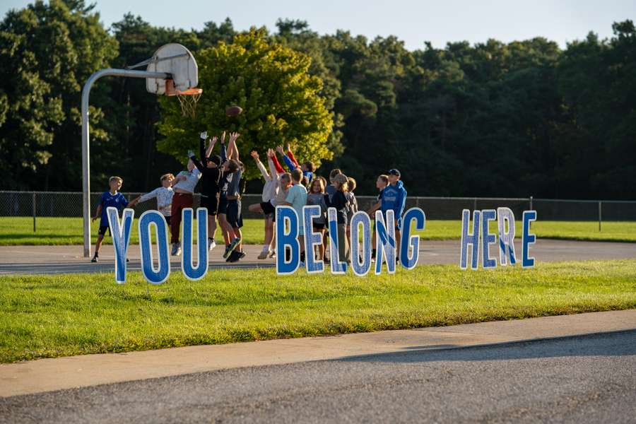 A group of young students leaping up to catch a football on the playground, behind letters in the foreground that spell out YOU BELONG HERE