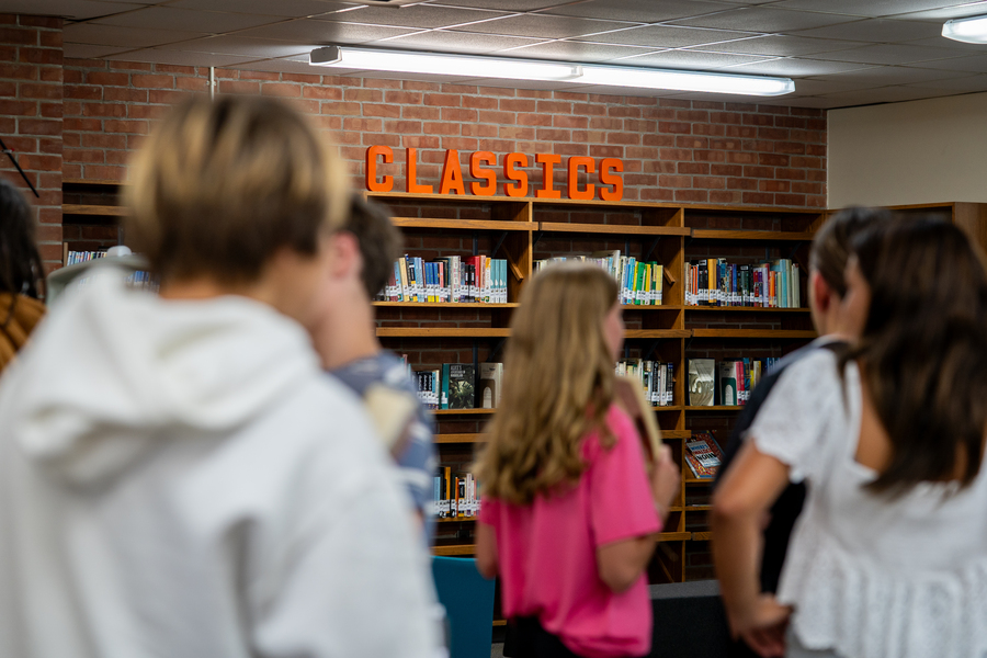 Blurred out students in the foreground, standing in a library, with a bookshelf behind them that has a sign saying CLASSICS on top of it.