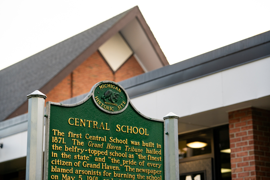 A close-up of the “Central School” historic marker in front of a brick building with a peaked roof. The top of the sign features the Michigan Historic Site emblem. The marker text describes the origins and history of Central School in Grand Haven.